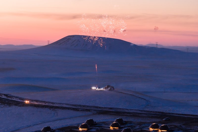 Cluster of dome-shaped shelters in a snowy field at dusk with illuminated volcano erupting in distance