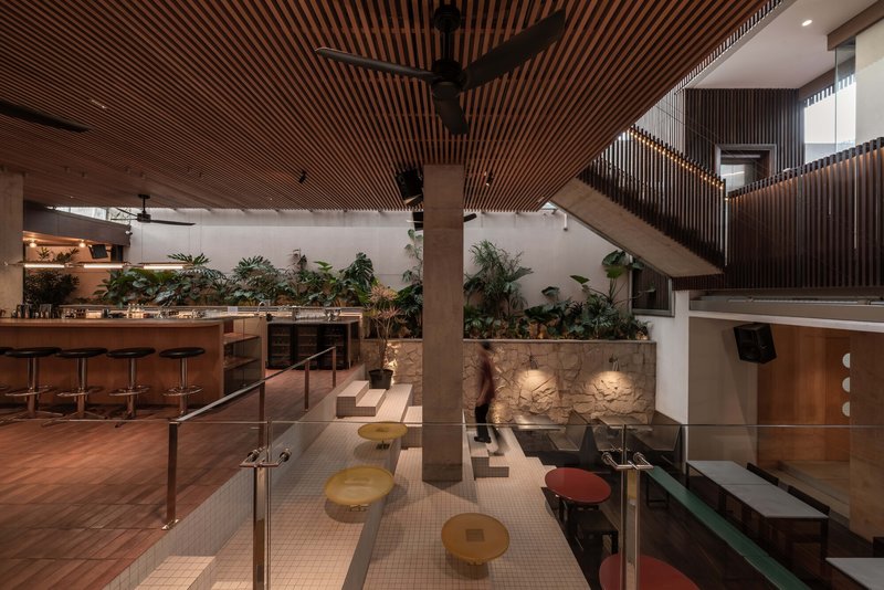 Double-height interior with timber-slatted ceiling, planted planter wall, and circular stools around glass-railed mezzanine