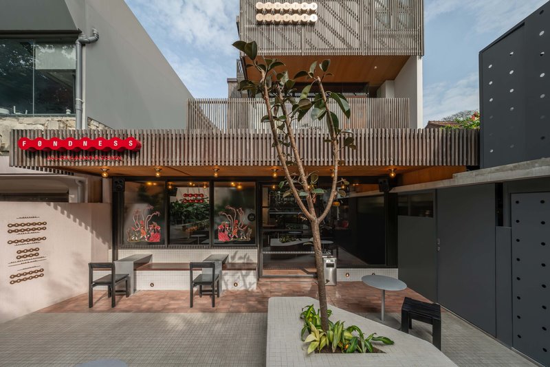 Ground-level courtyard with timber-screened upper volume, planted tree, and outdoor seating at dusk