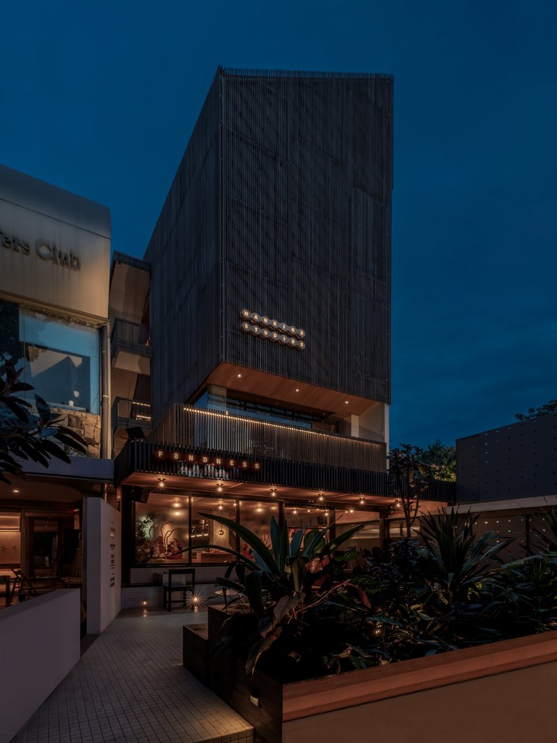 Street view of the dark timber-clad tower with illuminated signage and planted ground floor at twilight