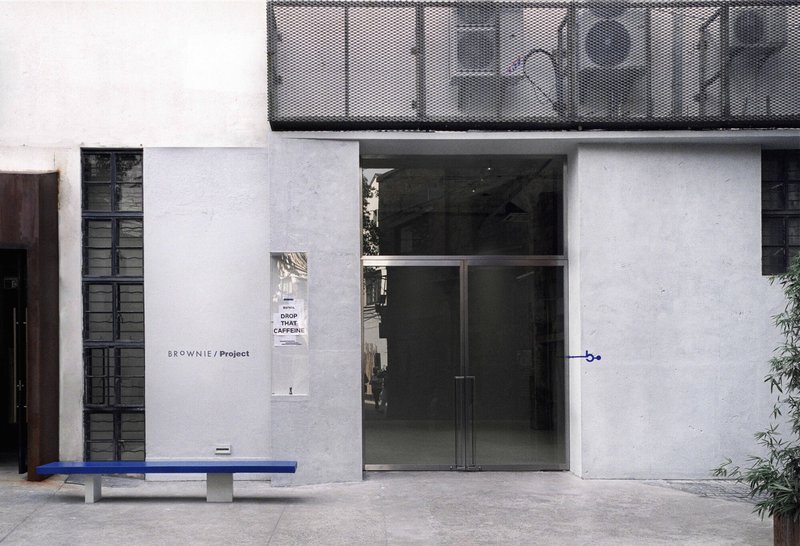 Street facade with glass entrance doors and perforated metal balcony above a blue painted bench