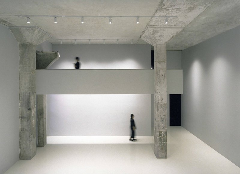 Double-height gallery space with illuminated horizontal display shelf and figure walking on upper level