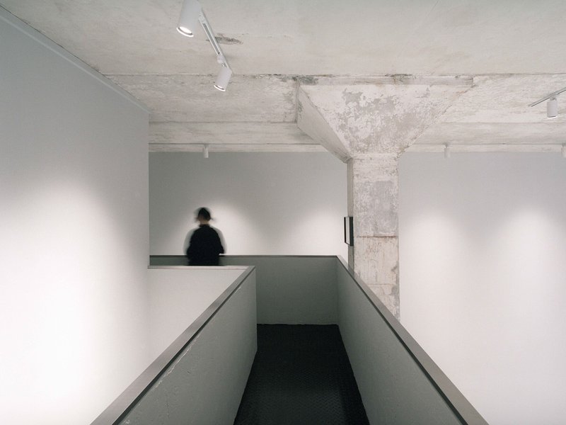 Ramp descending between white walls with concrete ceiling and figure visible on mezzanine beyond