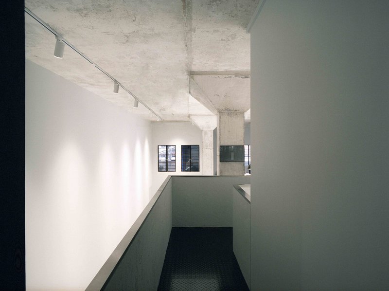 View from dark corridor across gray ramp balustrade toward lit white wall with window openings