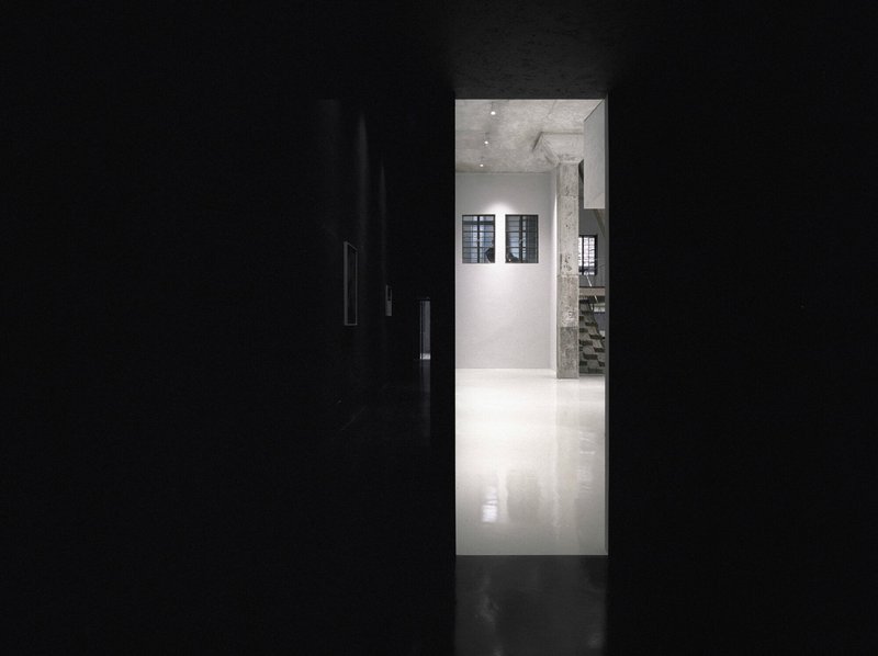 View through a dark corridor into a white-walled gallery space with concrete column and ceiling spotlights