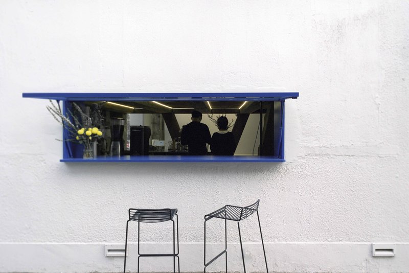 Blue-framed service window with mirrored interior set into white exterior wall above two wire stools
