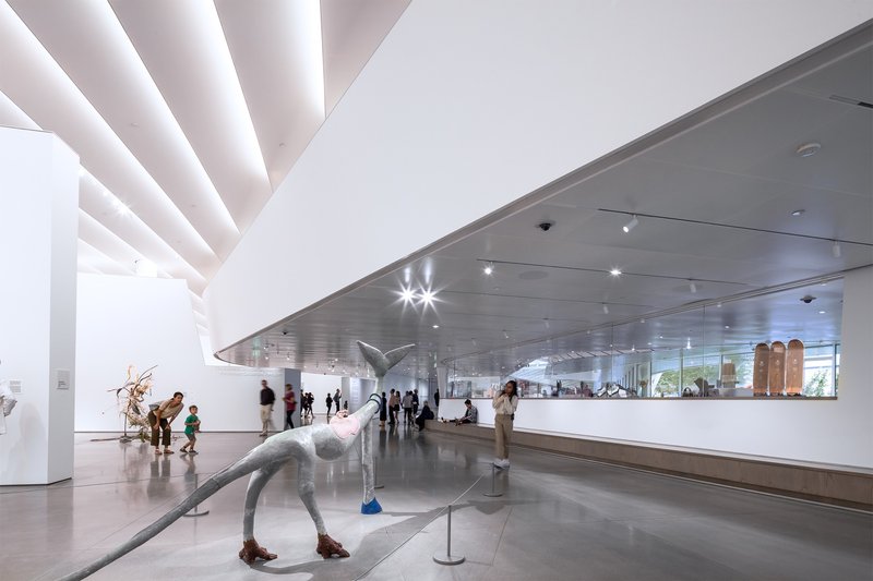 Gallery space with sculptural figures beneath a hovering metallic ceiling plane and skylights