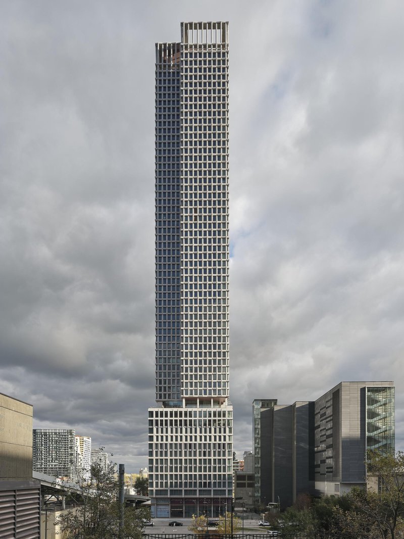 Slender residential tower with repeating grid facade rising above surrounding buildings under overcast skies