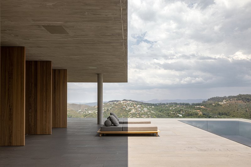 Covered terrace with infinity pool and low daybed facing hillside settlement under cloudy sky