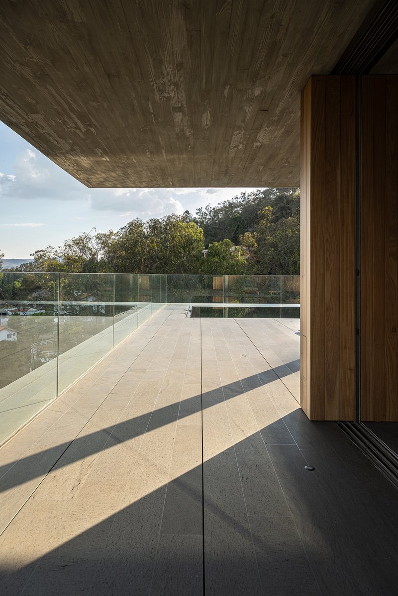 Glass-walled corridor with timber panels and stone flooring casting afternoon shadows across the terrace
