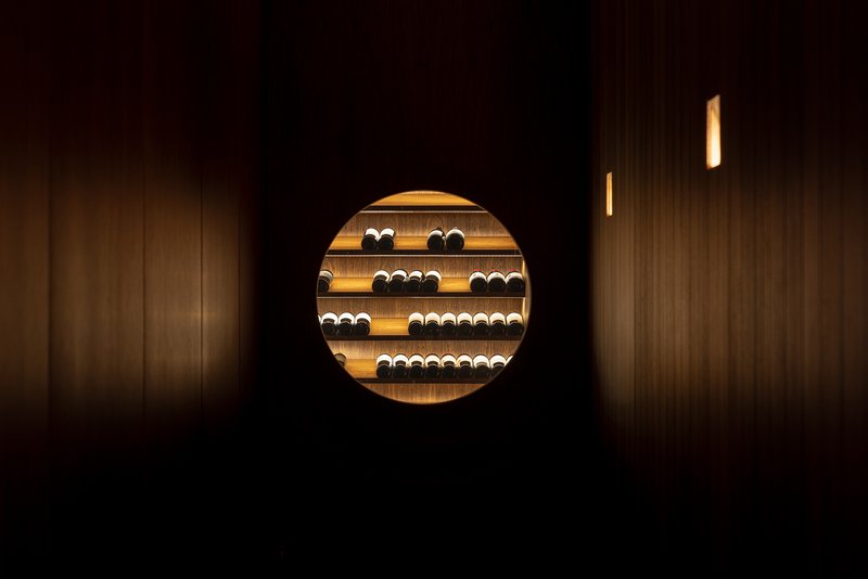 Circular aperture in a dark timber wall framing illuminated wine storage shelves