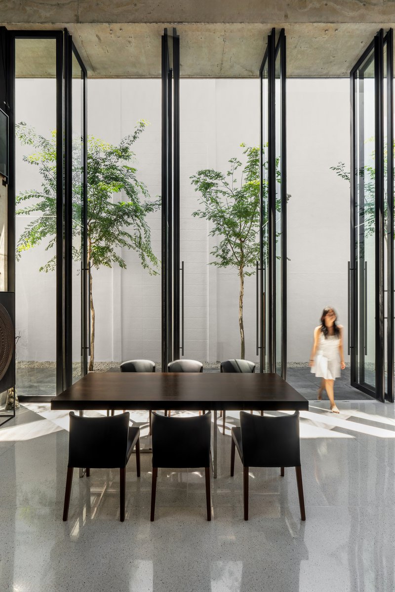 Black dining table beneath exposed concrete ceiling with tall glass walls framing courtyard trees and figure walking
