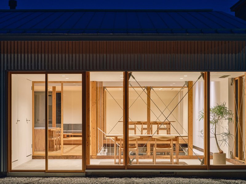 Glass wall revealing dining room with timber table and chairs beneath a corrugated metal roof at twilight