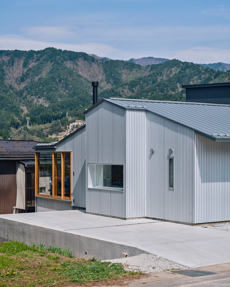 Gabled corrugated metal facade with punched openings and a concrete terrace against forested hills