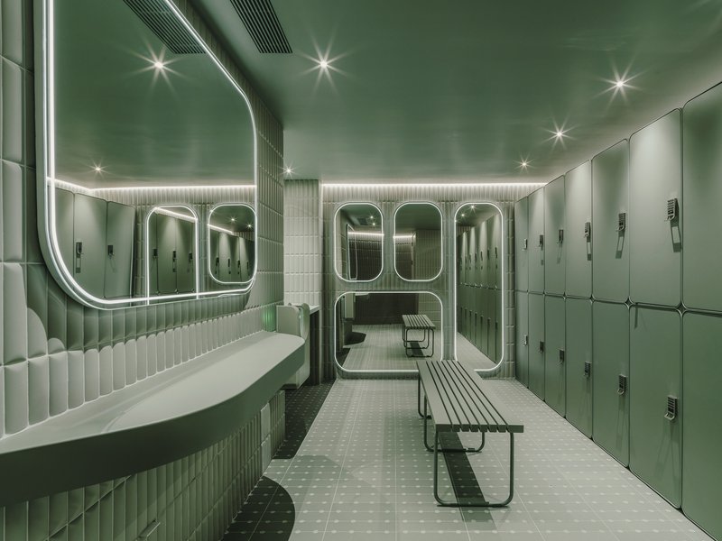 Locker room with fluted green tile walls, rounded mirrors with neon lighting and metal benches