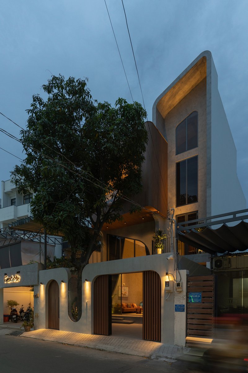 Street facade with arched openings and vertical timber slats illuminated at dusk beside a large tree