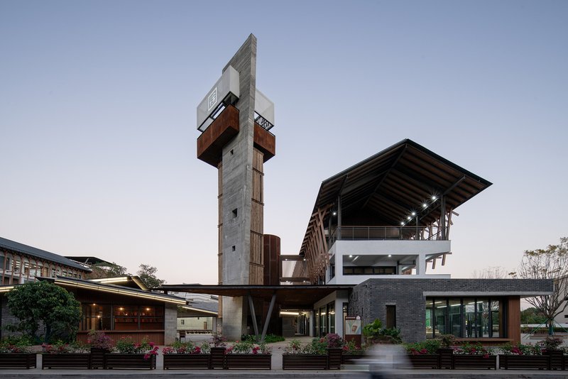 Concrete tower with cantilevered weathered steel volumes beside low-rise pavilions at dusk