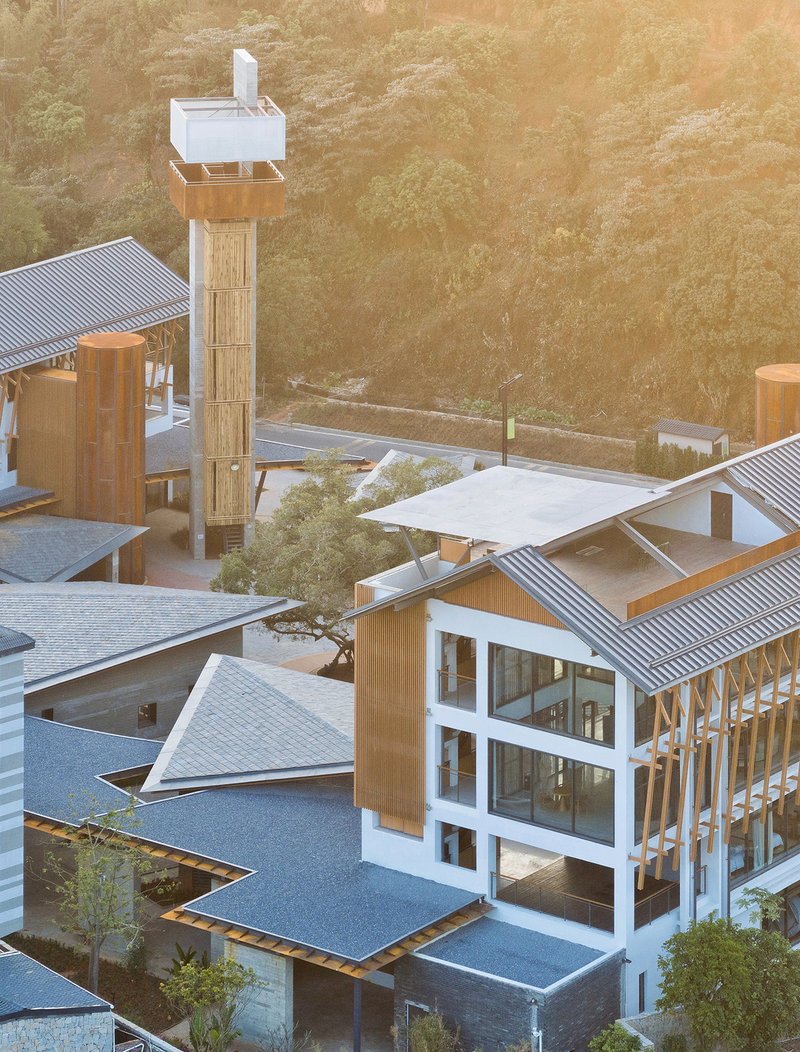 Aerial view of clustered volumes with corrugated metal roofs and timber-clad tower at golden hour