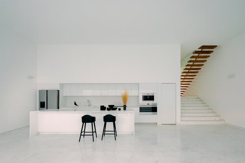 White kitchen island with black bar stools and floating timber staircase in a terrazzo-floored interior