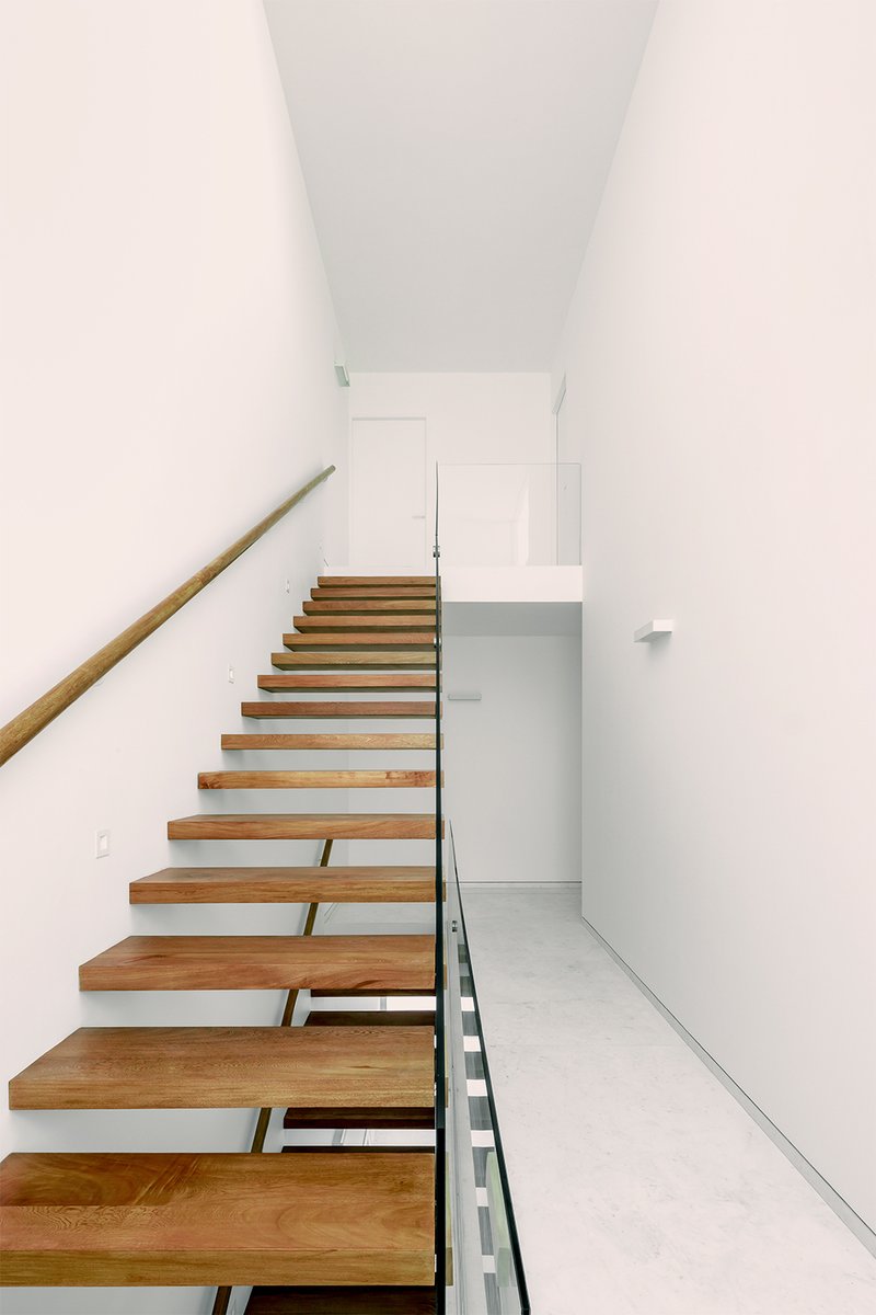 Open-riser timber staircase ascending through a narrow white-walled volume with wooden handrail