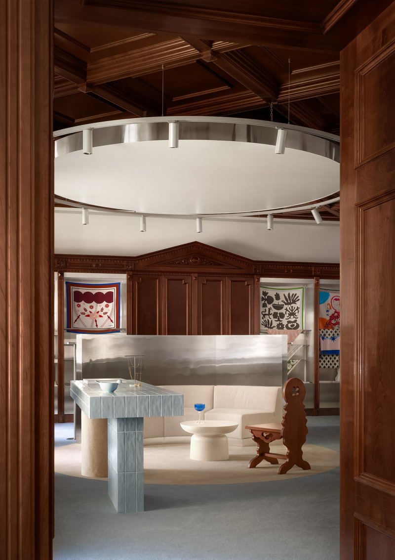View through doorway to circular suspended lighting fixture above ribbed display tables and coffered ceiling