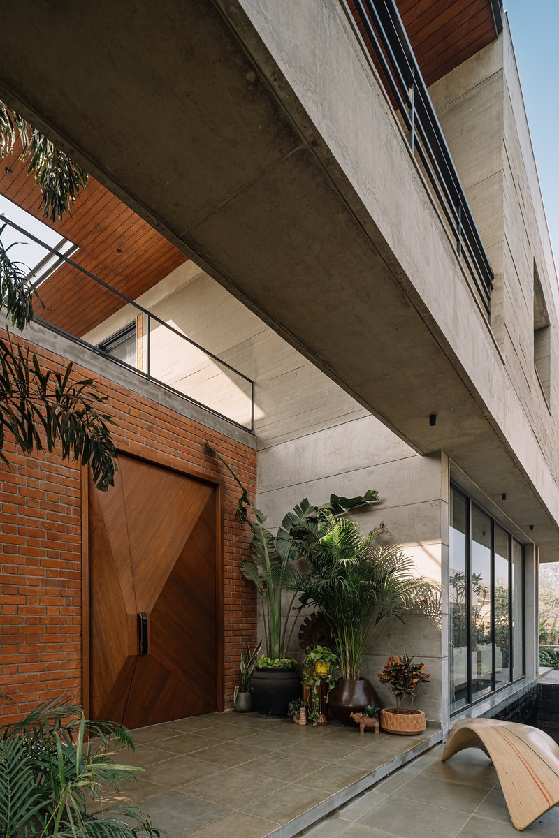 Covered entry courtyard with wooden pivot door, concrete soffit, potted plants and glazed upper volume