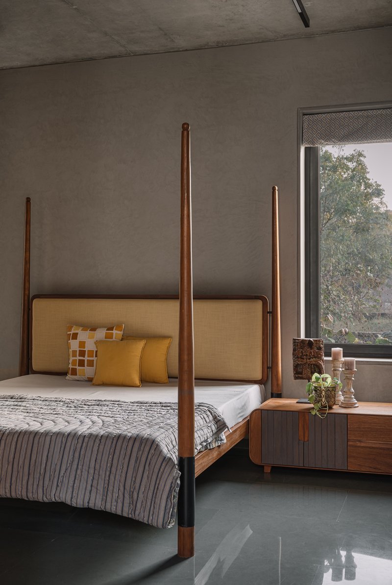 Four-poster bed with tall timber posts against a concrete wall with foliage visible through the window