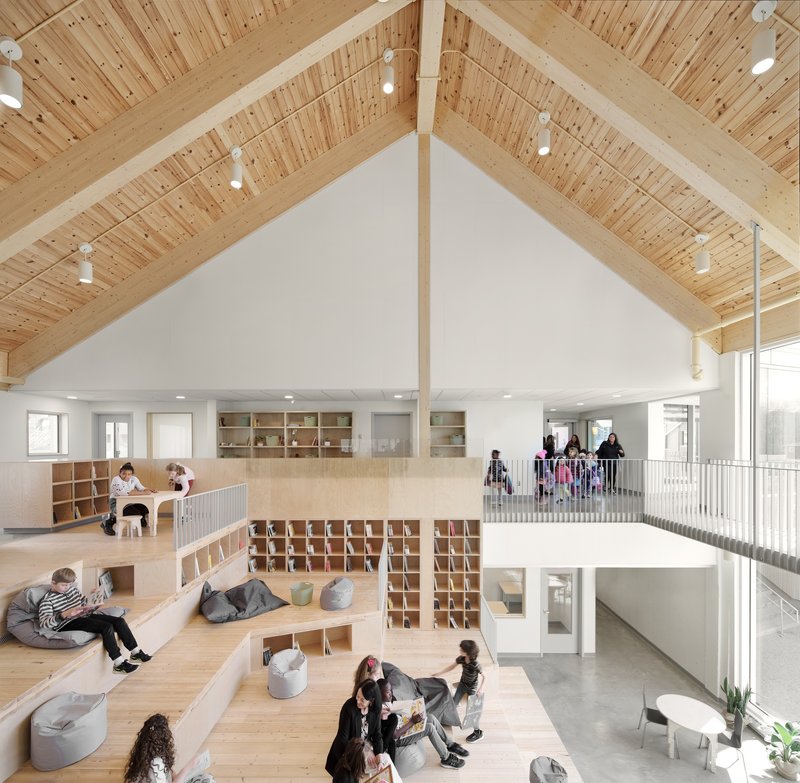 Double-height interior with exposed timber ceiling, plywood shelving, and children occupying tiered seating platforms