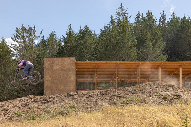 Cyclist jumping earthen berm beside rammed earth wall and timber-framed glazed facade in afternoon sun