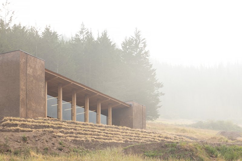 Timber-clad volume with exposed columns and glazing set into a hillside under dense fog