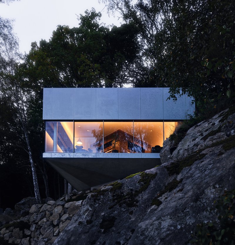 Cantilevered glass-walled volume with illuminated interior perched above a rocky slope at dusk