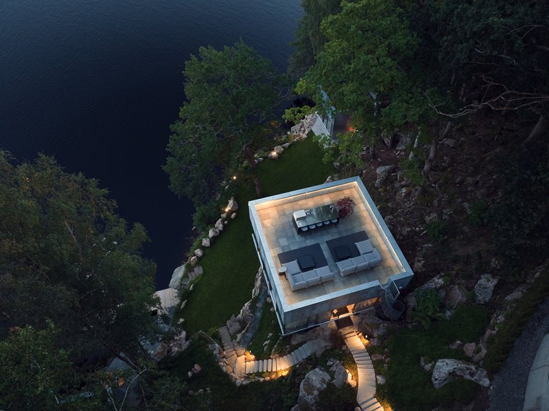 Aerial view of flat-roofed glass pavilion with terrace seating nestled among boulders beside dark water