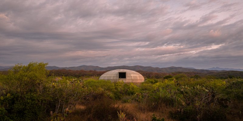 Domed structure with timber cladding and rectangular opening set among desert scrub under overcast skies