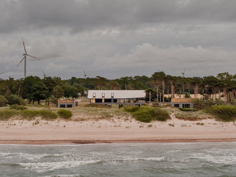 View from the beach showing the low-slung volumes with metal roof amid dune grasses and distant wind turbines