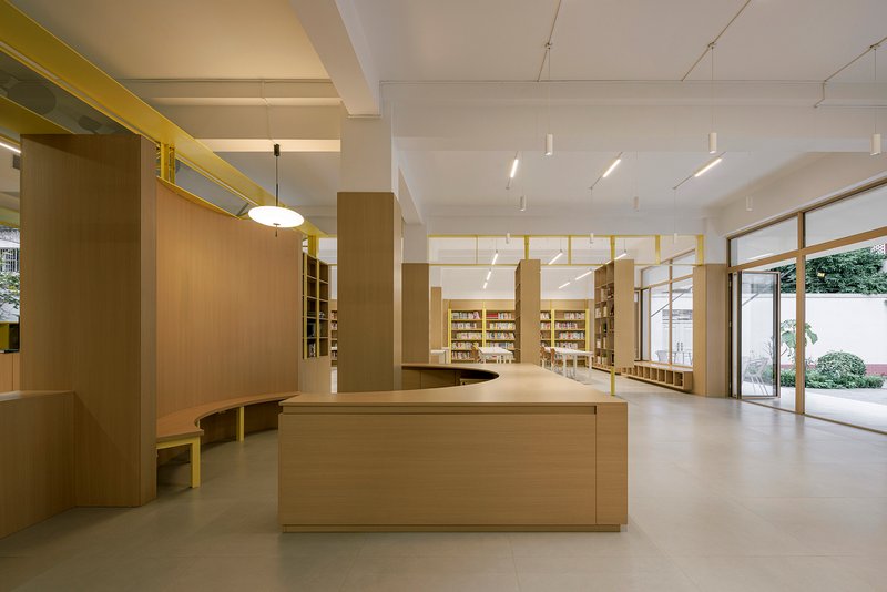 Central timber service counter beneath yellow ceiling beams with glazed doors opening to a courtyard beyond