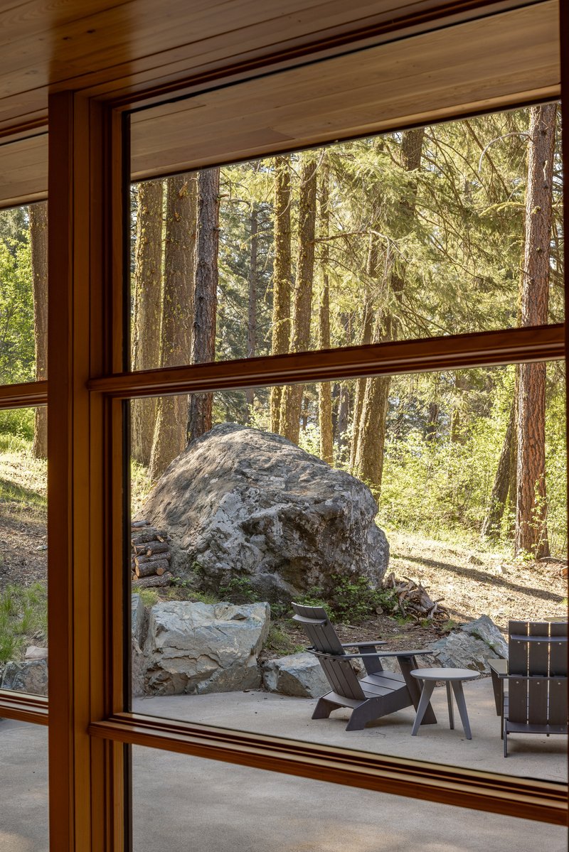 Timber-framed window looking out to a concrete patio with a large boulder and conifer trees beyond