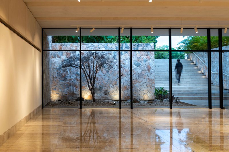 Interior gallery with polished stone floor and glazed wall revealing lit courtyard tree and exterior stair