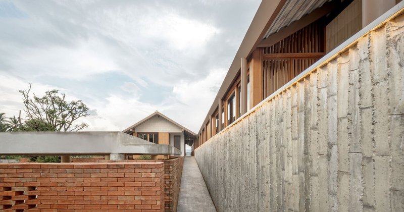 Narrow concrete pathway flanked by boardformed concrete wall and perforated brick wall beneath cloudy sky