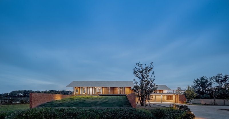 Low brick perimeter wall enclosing glass pavilions on a grassy berm at dusk