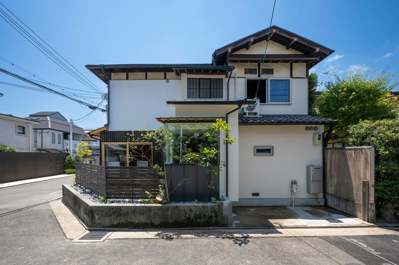 Street facade with white stucco walls, dark horizontal screens and small planted garden on a clear day