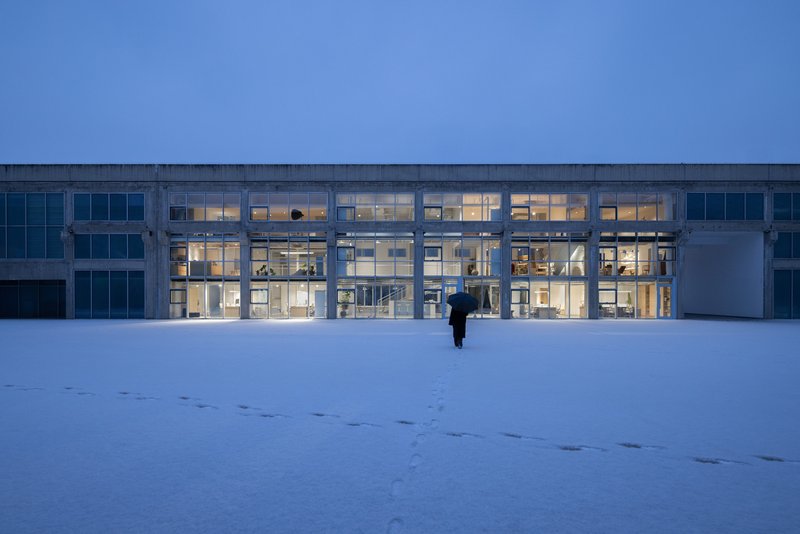 Illuminated glass facade with concrete structural frame viewed across a snow-covered field at dusk