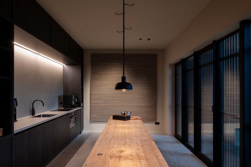 Galley kitchen with dark cabinetry, timber table and horizontal louvered screen illuminated at dusk