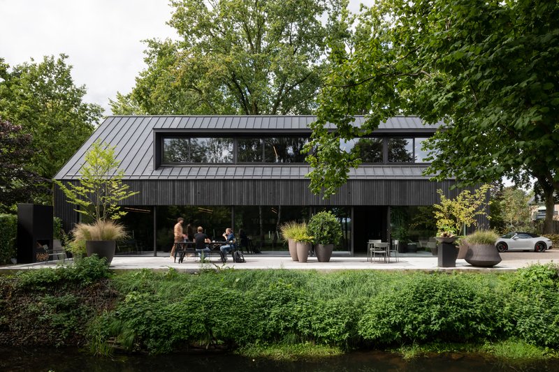Black timber-clad facade with standing seam metal roof and terrace occupied by people among grasses and planters