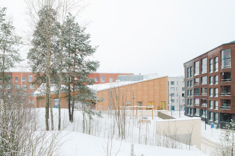 View across snow-covered terrain with pines toward the angled timber-clad volume and surrounding residential blocks