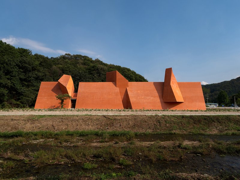 Long view of the angular orange brick volumes beside a forested hillside and plowed field
