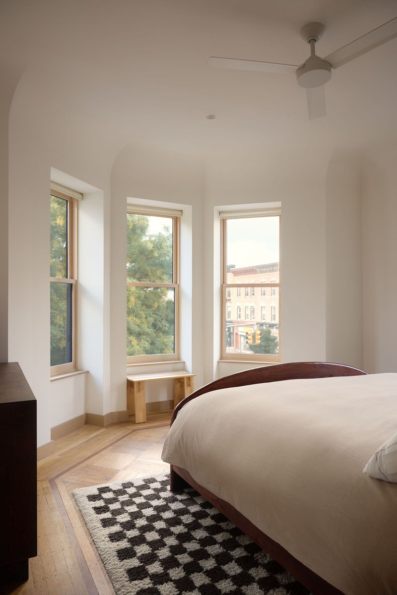 Bedroom with three tall windows framing views of neighboring buildings and surrounding trees