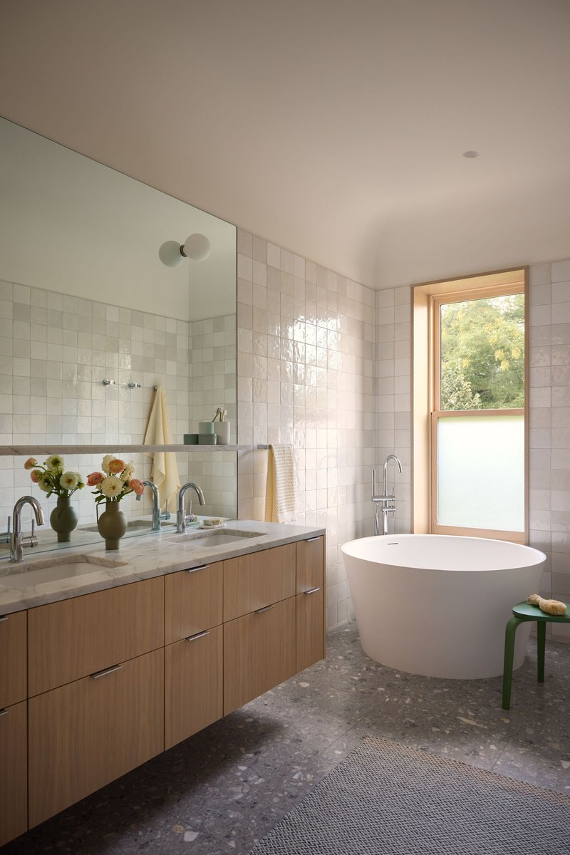 Bathroom with freestanding tub, oak vanity, white tile walls, and frosted window overlooking greenery