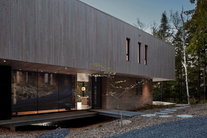 Weathered timber-clad facade with vertical slat windows and covered entry terrace beside a young tree in gravel