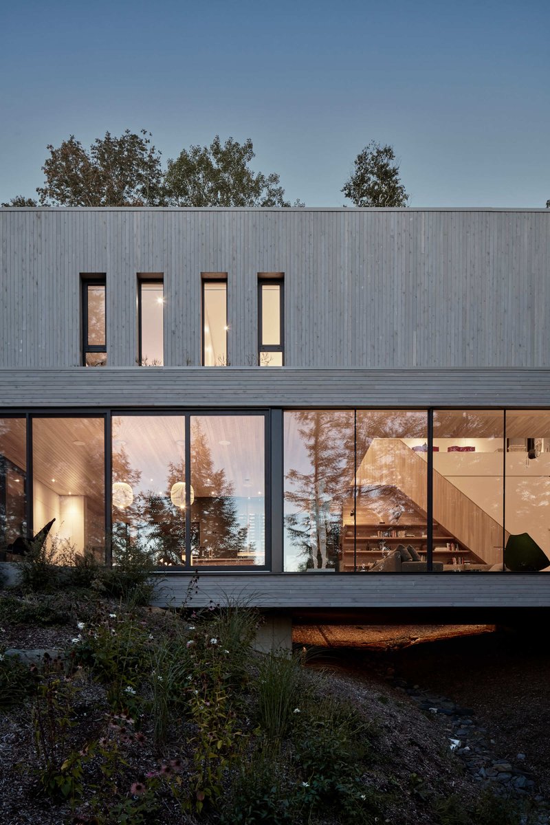 Two-story glazed facade with upper punched windows and lower glass wall reflecting surrounding pines at dusk