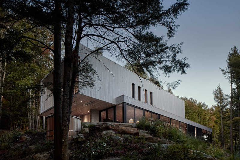 Two-story wood-clad volume on a rocky outcrop framed by tall pine trees at dusk
